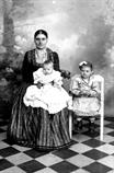 Ritratto di donna con due bambini, una seduta su una sedia e uno sulle ginocchia della donna. Alle spalle un fondale da studio fotografico